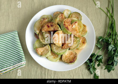 Simple healthy vegetarian food./ Many fried zucchini slices on round white plate and fresh green parsley over green fabric background top view closeup Stock Photo