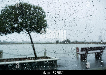 Raindrops on the windshield on a rainy day; bench on the shoreline of a lake in the background