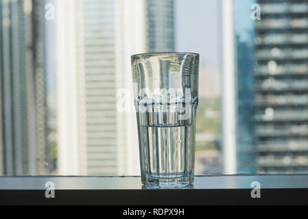 A half filled glass of water placed against window with skyline background. Clear space to write headline. Stock Photo