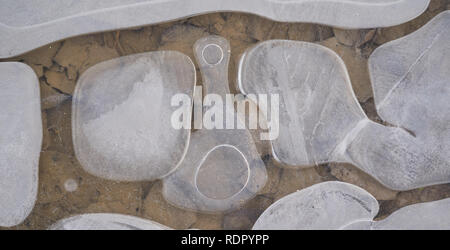 Abstract close-up of translucent ice pieces and trapped air bubbles over pebbles in shallow water, creating organic winter patterns Stock Photo