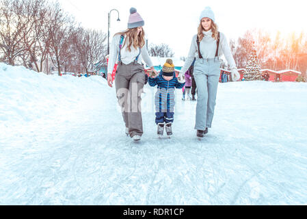 Two girls hold the hand of a little boy. In winter, city skate. Front view. Mom with a girlfriend and son. Stock Photo