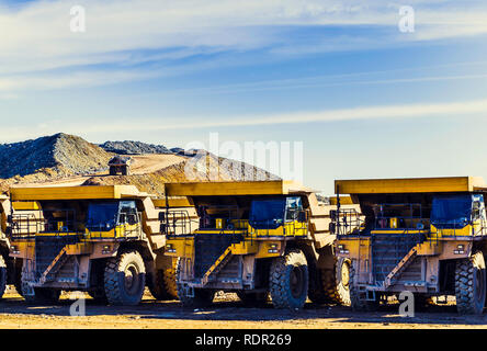 Big yellow dumper trucks placed in a row in the mine with dumper trucks laden with ore on the quarry road with blue sky and clouds in the background Stock Photo