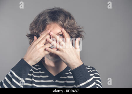 Handsome man with dark curly hair, bristle, hides face with hands, peeks through fingers, wears striped sweater, feels shy, isolated over grey backgro Stock Photo