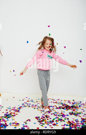 Happy beautiful little girl enjoying colorful confetti surprise falling down, posing on white studio wall. Pretty girl celebrating her birthday party, Stock Photo