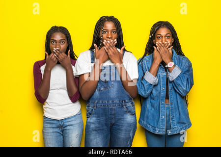 Portrait of three excited african women covering mouthes while telling secrets over yellow background Stock Photo