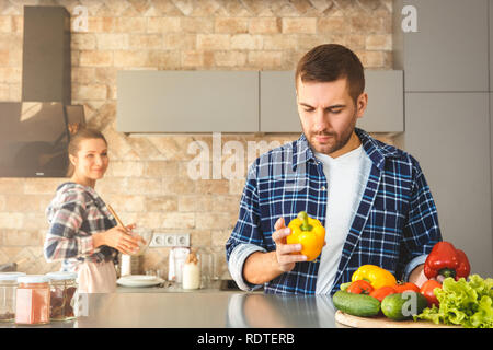 Young couple at home standing in kitchen together man holding bell pepper thoughtful while wife cooking joyful Stock Photo