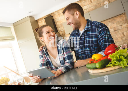 Young couple at home standing in kitchen together husband hugging wife using digital tablet looking at each other laughing Stock Photo