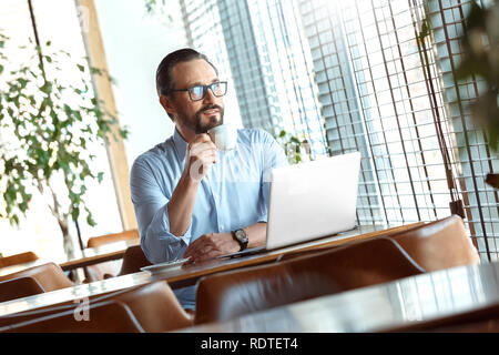 Business Lifestyle. Trader in glasses sitting at cafe with laptop drinking hot coffee enjoying view from the window smiling Stock Photo