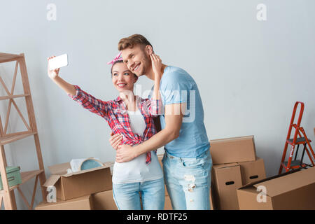 Young couple moving to new place standing hugging taking selfie photos with smartphone smiling joyful Stock Photo