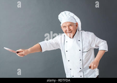 Senior chef studio standing isolated on gray playing with knives cheerful Stock Photo
