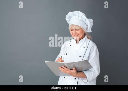 Senior woman chef studio standing isolated on gray writing in journal joyful Stock Photo
