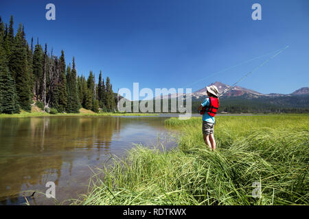 Girl fishing from shore along channel at Sparks Lake, Broken Top Mountain in background, Cascade Lakes, Oregon, USA Stock Photo