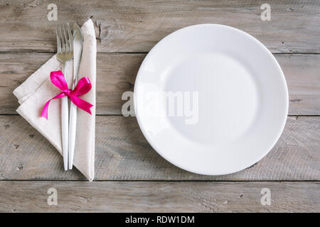Empty table setting with pink ribbon - plain white ceramic plate, linen napkin, cutlery on wooden table. Romantic date, dinner concept, nordic style. Stock Photo