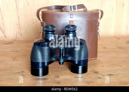 Vintage Porro prism black color binoculars and closed brown hard leather case on wooden background front view indoor closeup Stock Photo