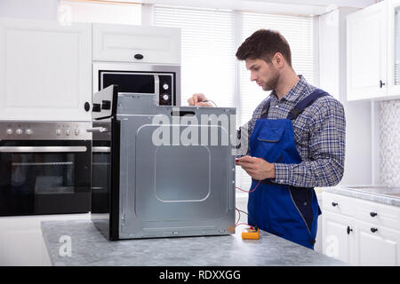 Young Male Repairman Repairing Oven Using Digital Multimeter In Kitchen Stock Photo
