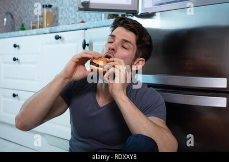 Close-up Of A Man Eating Burger Leaning On Refrigerator Stock Photo