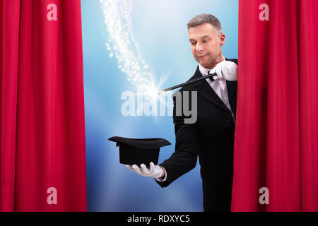 Magician Doing Magic Trick With Illuminated Wand And Hat Behind Curtain Stock Photo