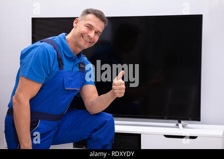 Smiling  Mature Technician Showing Thumb Up In Front Of Television Stock Photo