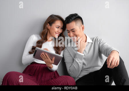 Happy young couple using tablet sitting on the floor at home isolated on gray background Stock Photo