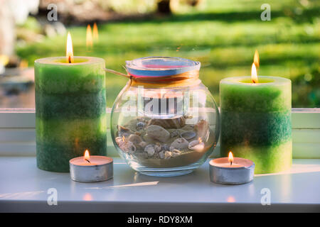 Multiple candles including votive candles, large green candles, and a te light in a bowl with shells are burning on a windowsill Stock Photo