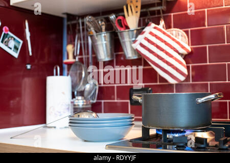 grey cooking pot on gas stove with steam releasing and blue dishes on the side in red burgundy classic kitchen with tools in the background. Stock Photo