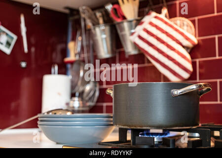 grey cooking pot on gas stove with steam releasing and blue dishes on the side in red burgundy classic kitchen with tools in the background. Stock Photo