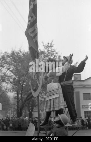 It was demonstration on 1-st May of 1978. Motorcyclist carries symbol of the Soviet era-the worker and the peasant. They stand upright on pedestal made directly on sidecar. The man and woman are specially dressed in appropriate clothes. They hold the flag of the USSR and metal hammer and sickle. The motorcyclist rides ahead of long column. The procession passes through the Central square of small town. Citizens with a sinking heart are look at the living symbol of communism. Stock Photo