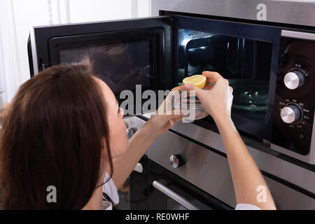 Woman Putting Sliced Lemon In Bowl Near Open Microwave Oven Stock Photo