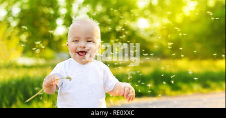 Funny child laughing. Portrait smiling happy baby boy playing spring, summer day,outdoors. Emotion face kid,Close up. Dandelion seeds blowing in wind across natural background park, banner,copy space Stock Photo