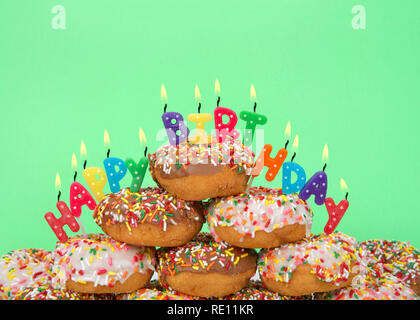 Chocolate and white frosted donuts covered in candy sprinkles piled into a cake pile with Happy Birthday candles burning brightly. Green background. Stock Photo