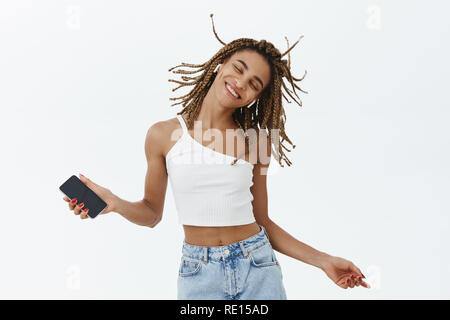 Music making life colourful. Portrait of carefree charming dark-skinned girl with dreadlocks jumping and dancing smiling with closed eyes having fun w Stock Photo