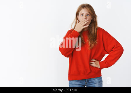 Studio shot of surprised and wondered attractive young slender good-looking woman holding hand pressed to mouth, gasping looking shocked and amazed at Stock Photo