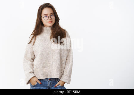 Warm winter days in cozy outfit. Portrait of charming smart and confident brunette in glasses wearing sweater, holding hands in pockets smiling cute a Stock Photo
