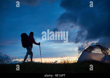 Young female climber reaching the top with walking sticks and tourist backpack at sunset in mountains in Romania. Night evening tent meadow slope landscape beautiful breathtaking scenery view nature Stock Photo