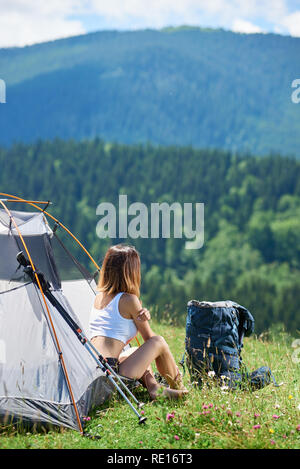 Back view of sporty young female climber sitting at tent entrance beside backpack and trekking sticks, enjoying sunny morning in the mountains. Lifestyle concept adventure summer vacations outdoor Stock Photo