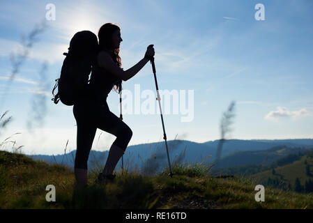 Silhouette of active girl climber with backpack and trekking poles climbing up on the top of a hill, agains blue sky in the mountains. Copy space. Concept of active lifestyle Stock Photo