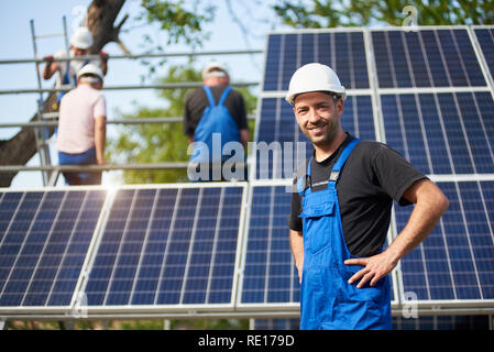 Portrait of smiling successful engineer technician standing in front of unfinished high exterior solar panel photo voltaic system blue shiny surface with team of workers on high platform. Stock Photo