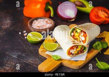 Grilled burritos wraps with chicken, beans, corn, tomatoes and avocado on a wooden board, dark background. Meat burrito, mexican food. Stock Photo