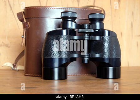 Vintage Porro prism black color binoculars and closed brown hard leather case on wooden background front view indoor closeup Stock Photo