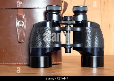 Vintage Porro prism black color binoculars and closed brown hard leather case on wooden background front view indoor closeup Stock Photo