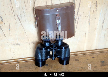 Vintage Porro prism black color army binoculars and closed brown hard leather carry case hangs on the wall front view indoor closeup Stock Photo