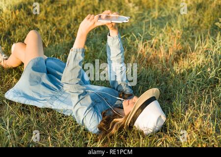 Portrait of teenage girl 14 years old lying on the grass. Girl in dress hat, in her headphones holds a smartphone, listens to music makes a selfi phot Stock Photo