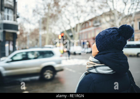 Unrecognizable young woman wearing winter knitted hat looking at road while standing on city street near crosswalk waiting for green traffic light. Stock Photo