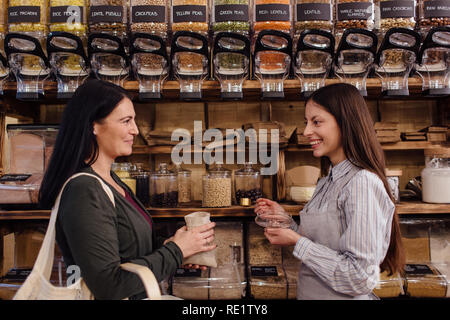 Smiling shop assistant and customer standing against each other in package free grocery store. Zero waste shopping -  shopkeeper serving customer. Stock Photo
