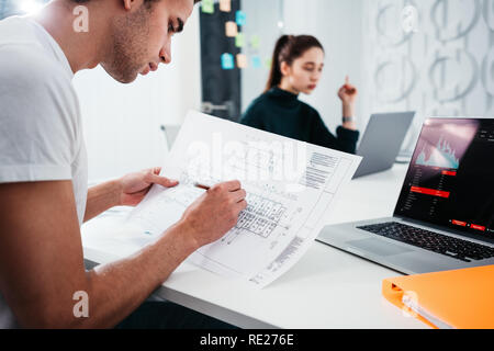 Young male architect preparing building blueprint plan sits at table and uses laptop. Team of engineer and designer develop building project Stock Photo