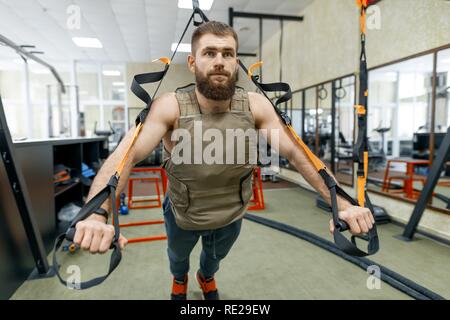 Muscular bearded man dressed in military weighted armored vest doing exercises using straps systems in the gym. Sport, training, bodybuilding and heal Stock Photo