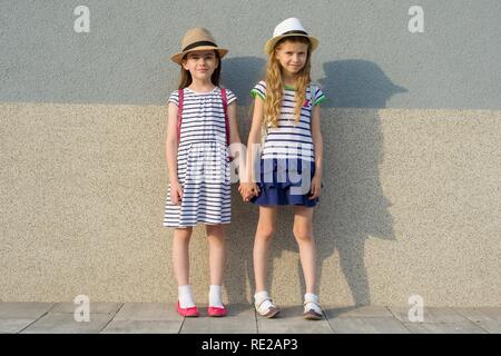 Outdoor summer portrait of two happy girl friends 7, 8 years holding hands. Girls in striped dresses, hats with backpack, background gray wall Stock Photo
