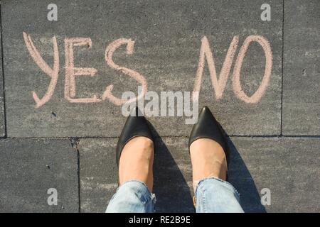 Woman before the choice yes or no. View from above, female feet with text yes no written on grey sidewalk Stock Photo
