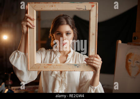 Female painter holds wooden frame at her face in studio, portrait drawing concept. Artist painting in class, workshop interior on background Stock Photo
