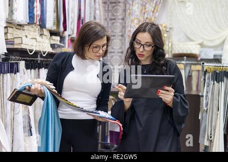 Female salesman and buyer in the fabric store are looking to select samples of materials. Stock Photo